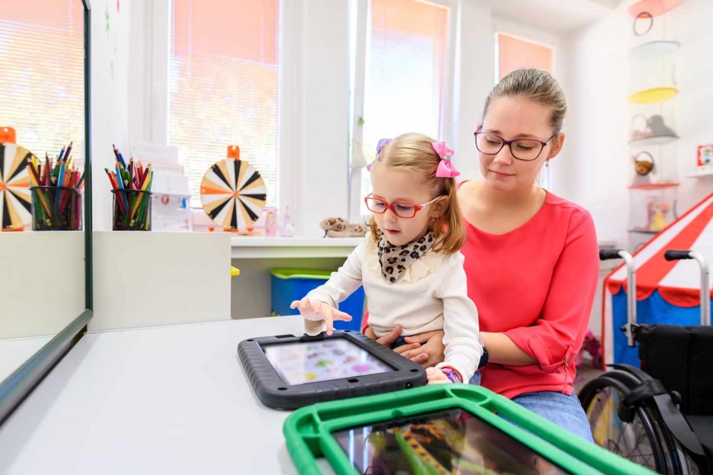 Mädchen mit Brille tippt auf einen Talker, eine Logopädin hält es auf dem Schoß