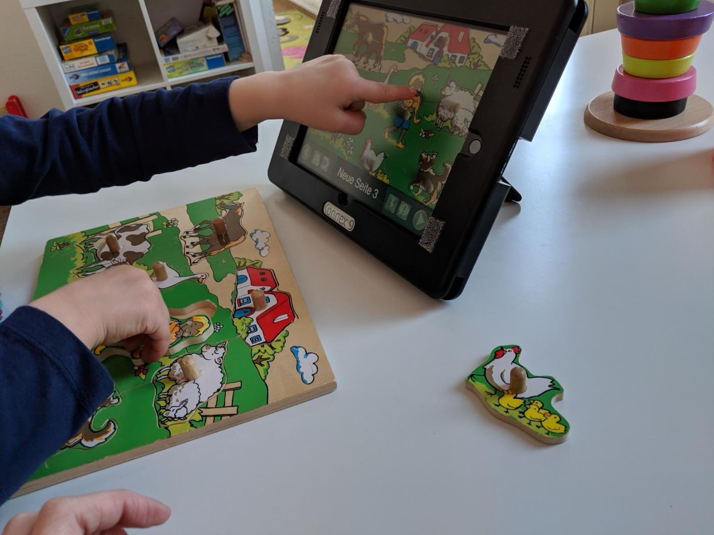 Kinderhand zeigt auf einem Talker auf ein Bild, daneben ein Holzpuzzle mit Bauernhoftieren