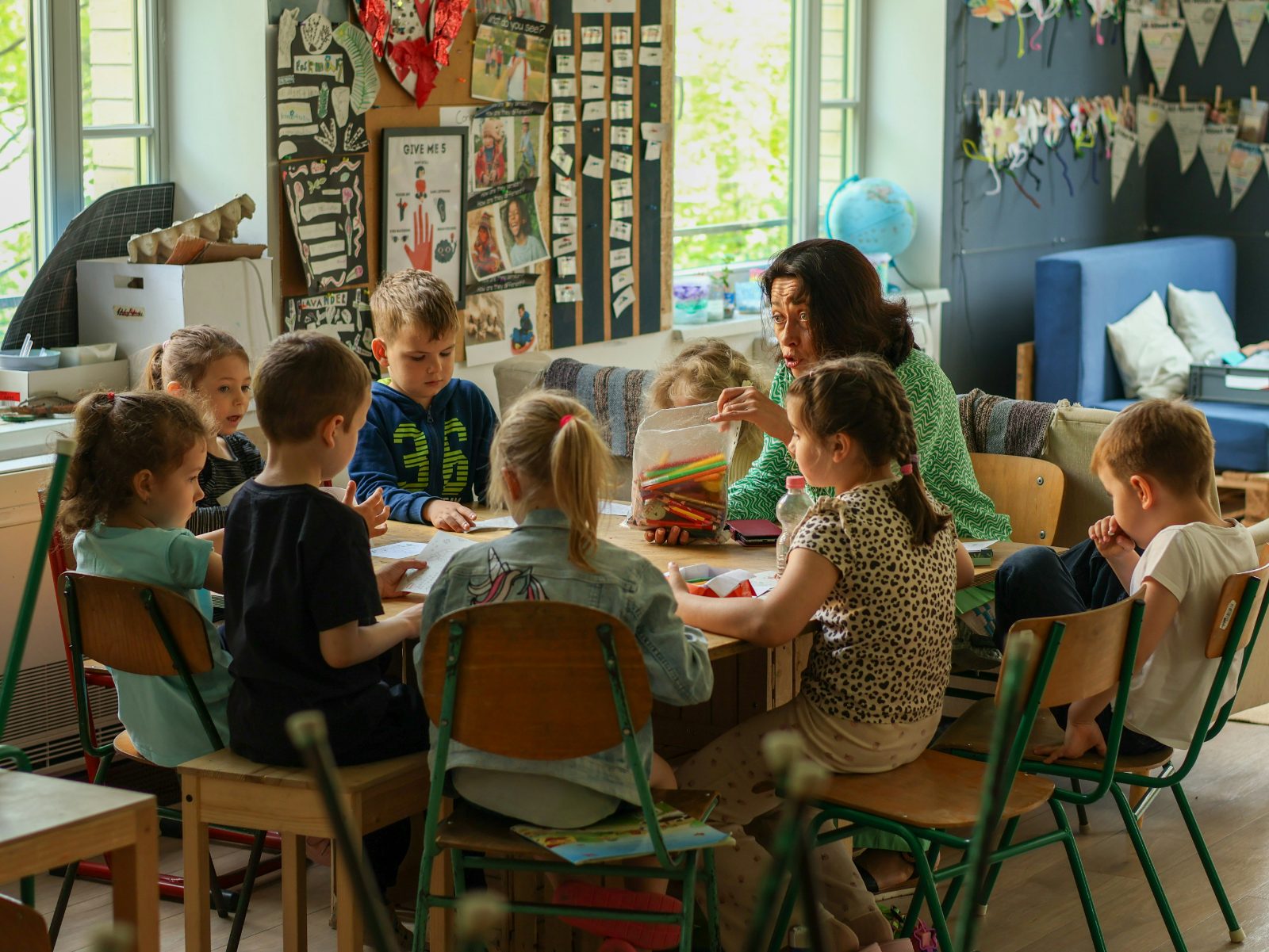 Erzieherin sitzt mit einer Gruppe Kita-Kinder am Tisch und spricht mit ihnen