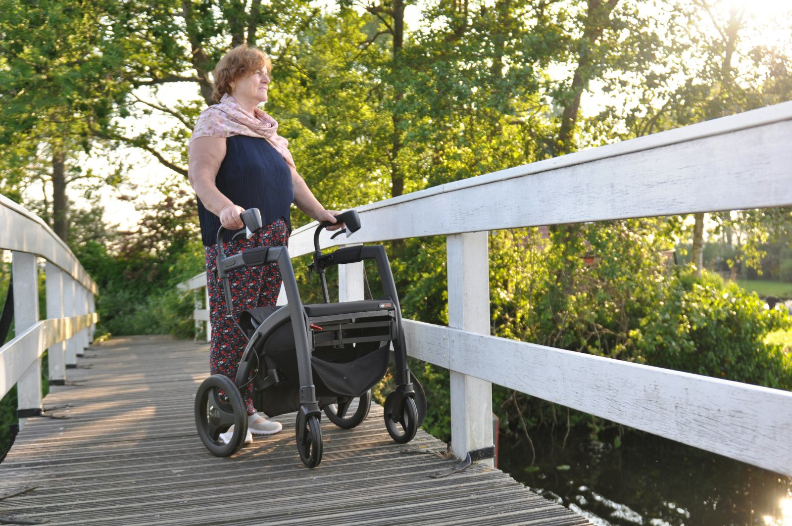 Ältere Frau mit Rollator steht auf einer Holzbrücke im Grünen